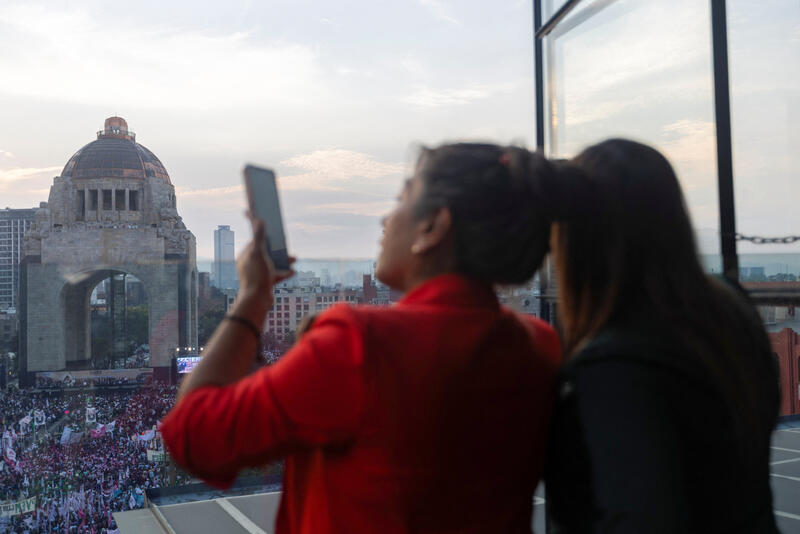 Two women in the foreground, one of whom is holding up a mobile phone, look down at a rally below.