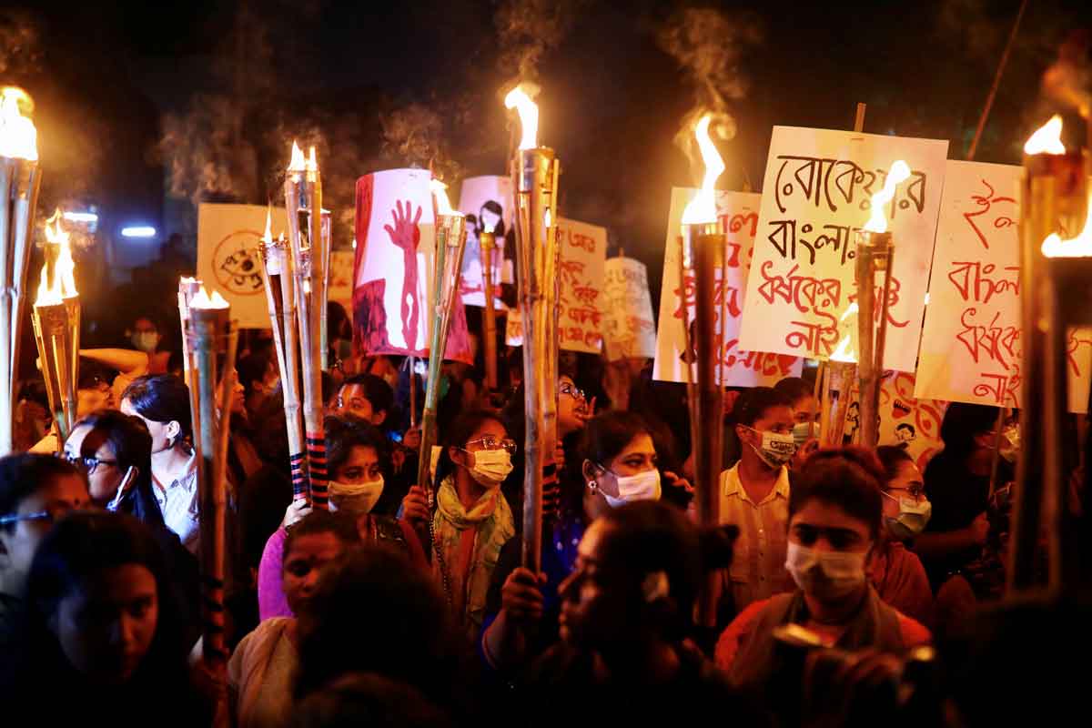 A crowd of protestors marching at night, carrying placards in Bangla script and torches