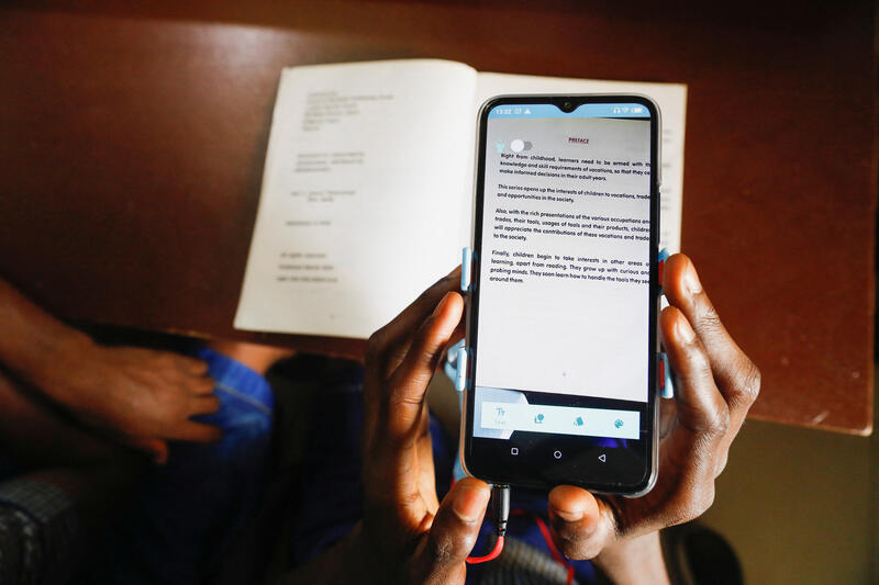 Hands holding up a mobile phone in front of a book. On the phone screen, we see enlarged text from the book.