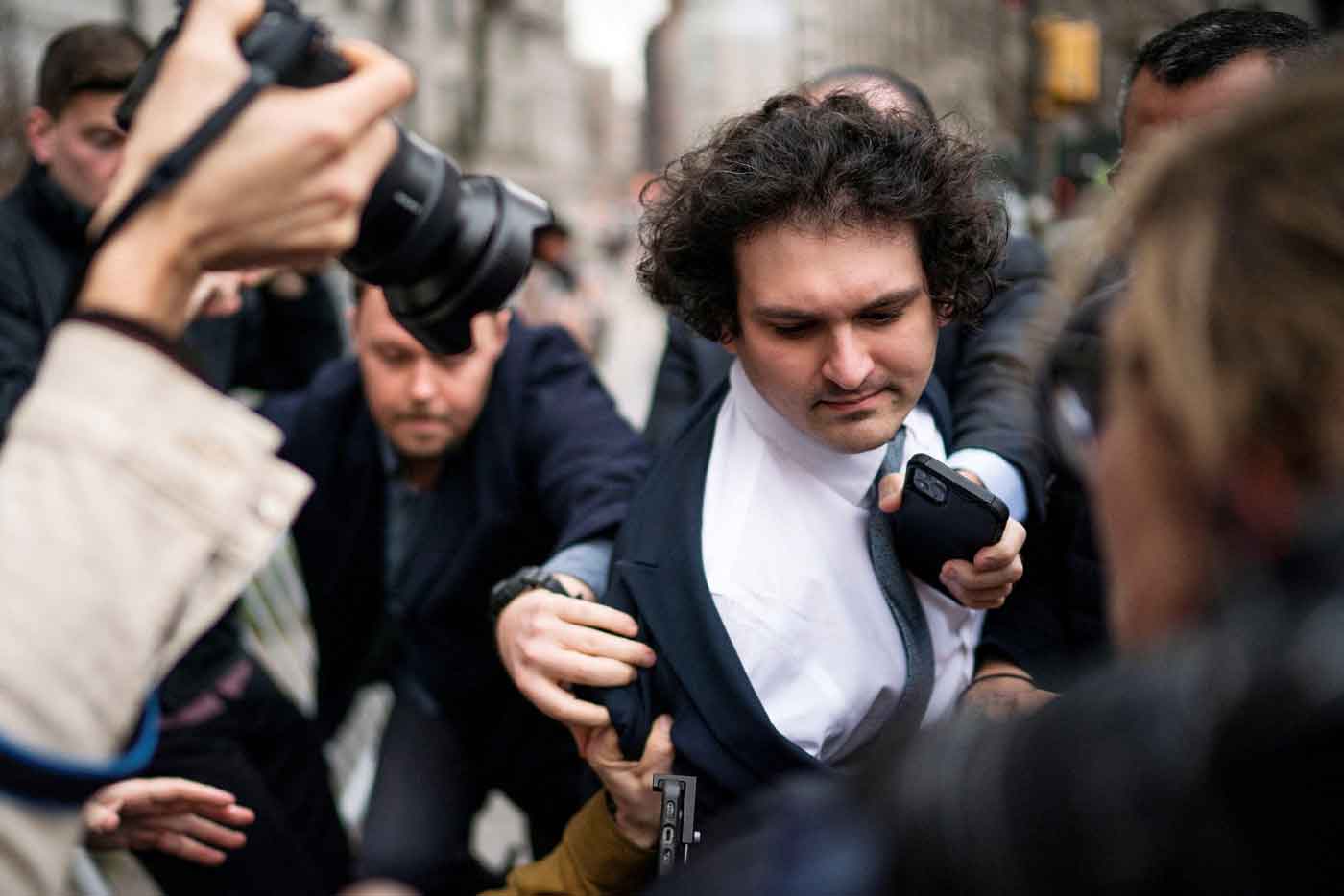 Curly headed man is jostled by a crowd of reporters with cameras and microphones