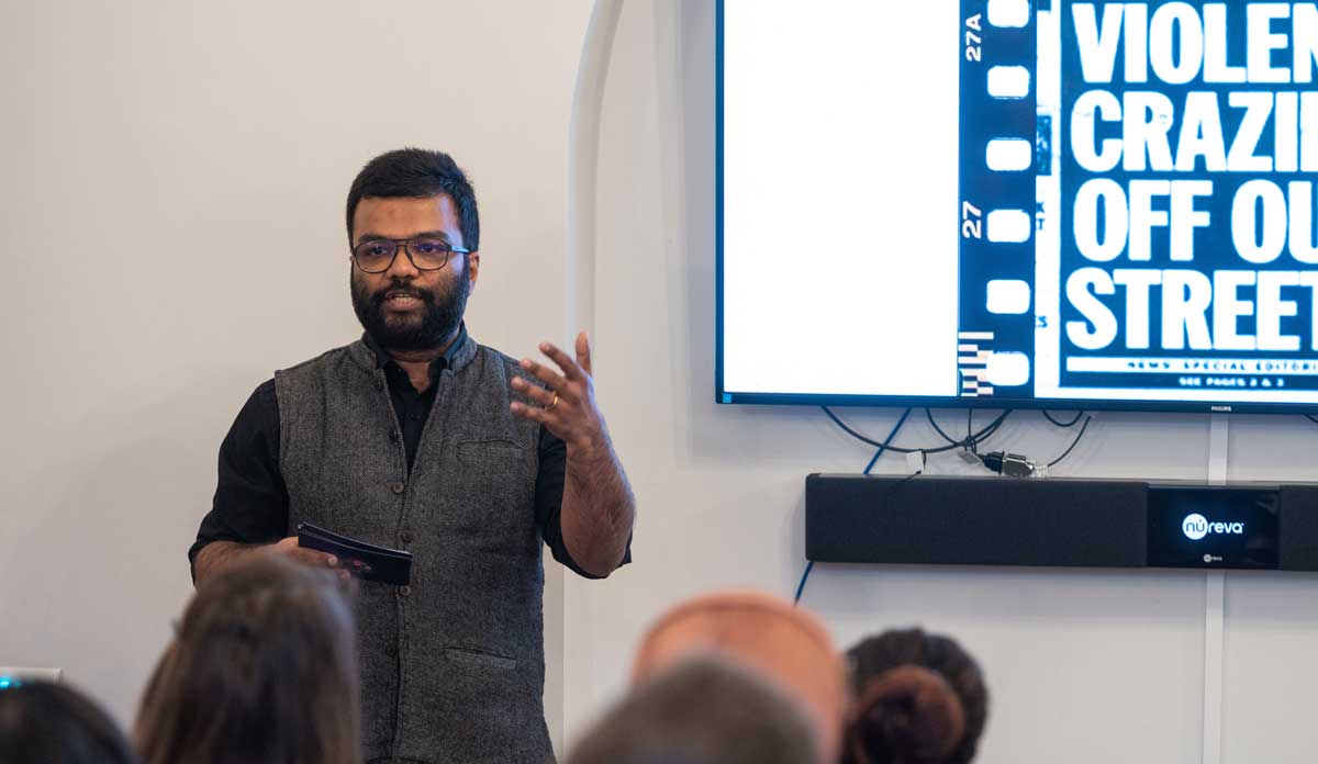 Tanmoy stands at the front of a room gesticulating. On the screen behind him is a tabloid newspaper headline that uses derogatory language about mental health disorders.