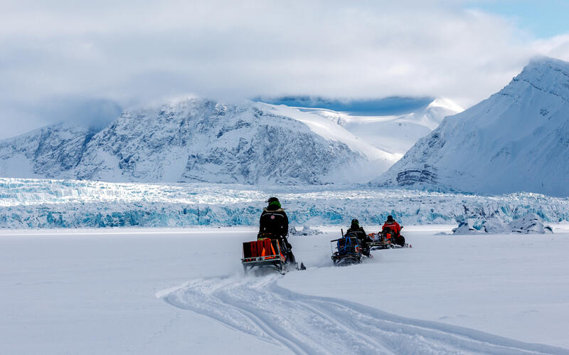 Three people on snowmobiles driving through a snowy landscape.