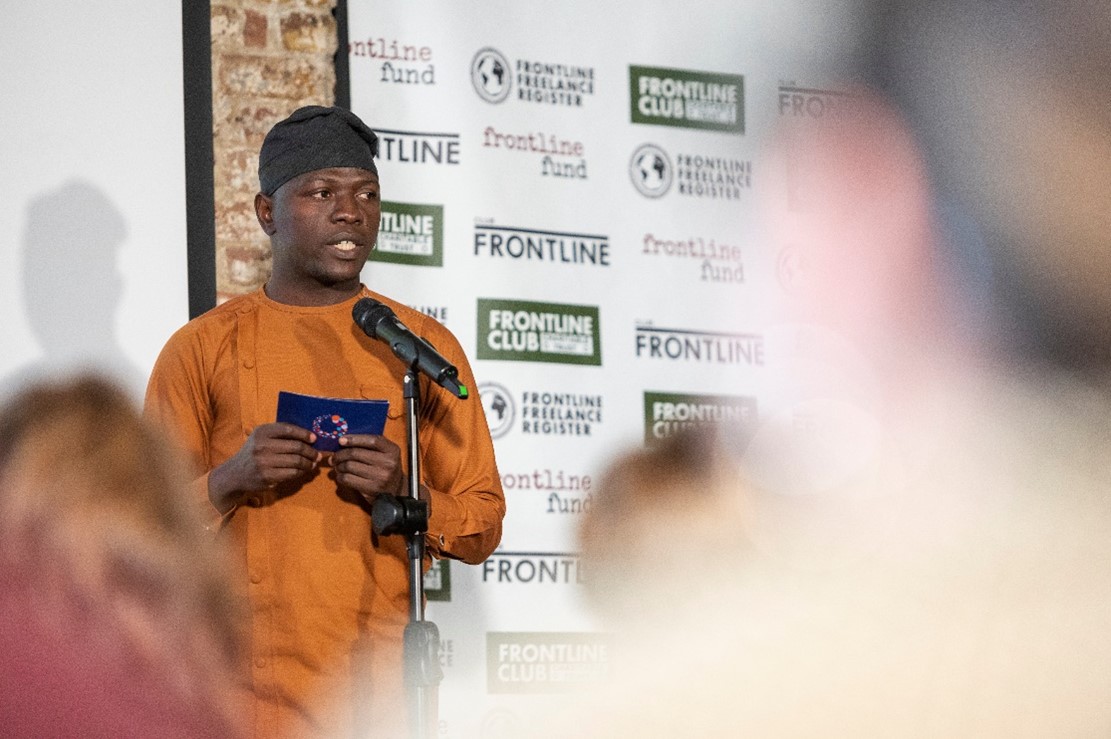 Samuel Thomas on stage at the Frontline Club in a burnt-orange Agbada (traditional Nigerian celebration attire)