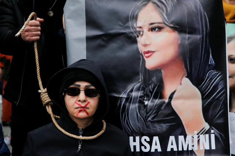 A woman in a black hoodie with sunglasses protests in front of a picture of Mahsa Amini. She has a red cross over her lips and a noose around her neck to signify the repression of women.