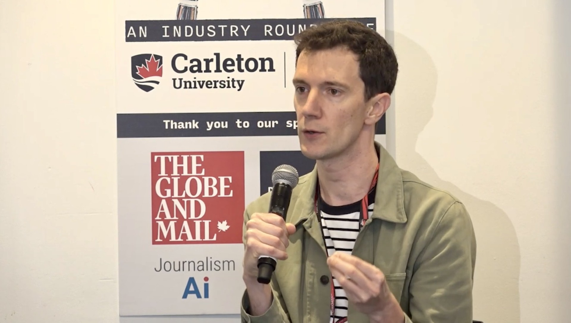 Florent Daudens speaks holding a microphone and gesticulating in front of a sign displaying the logos of several news and AI organisations.