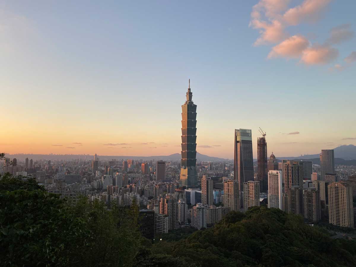 A panoramic view of Taipei at sunset, featuring Taipei 101 towering over the city's skyline. The sky transitions from soft blue to warm orange hues, with scattered clouds reflecting the evening light.