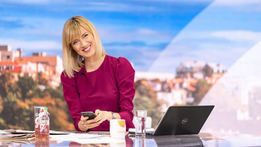 A woman with shoulder-length blonde hair wearing a fuchsia dress sits at a news desk, smiling warmly while holding a smartphone. 