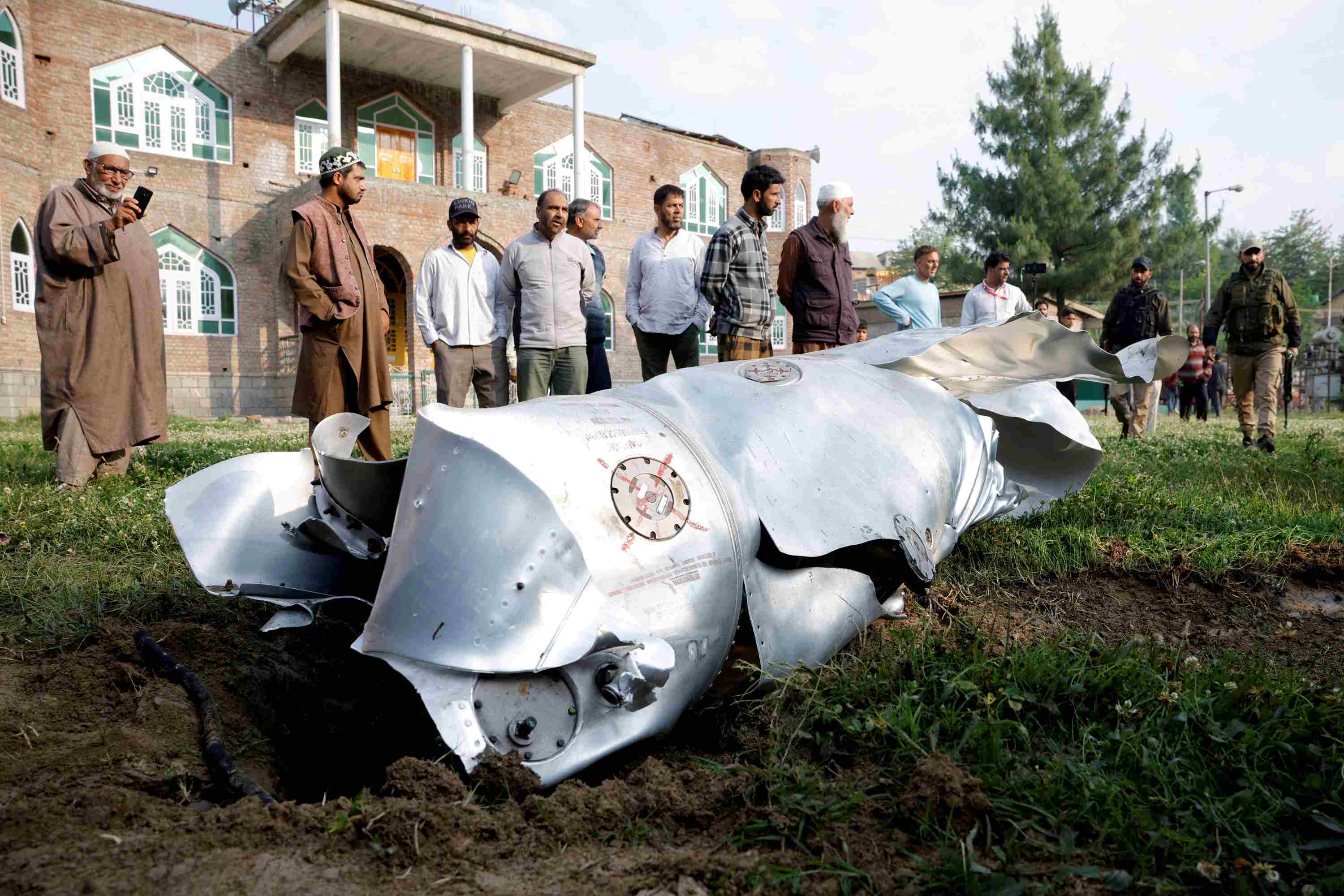 Metal debris lies on the ground, a group of men look at it.