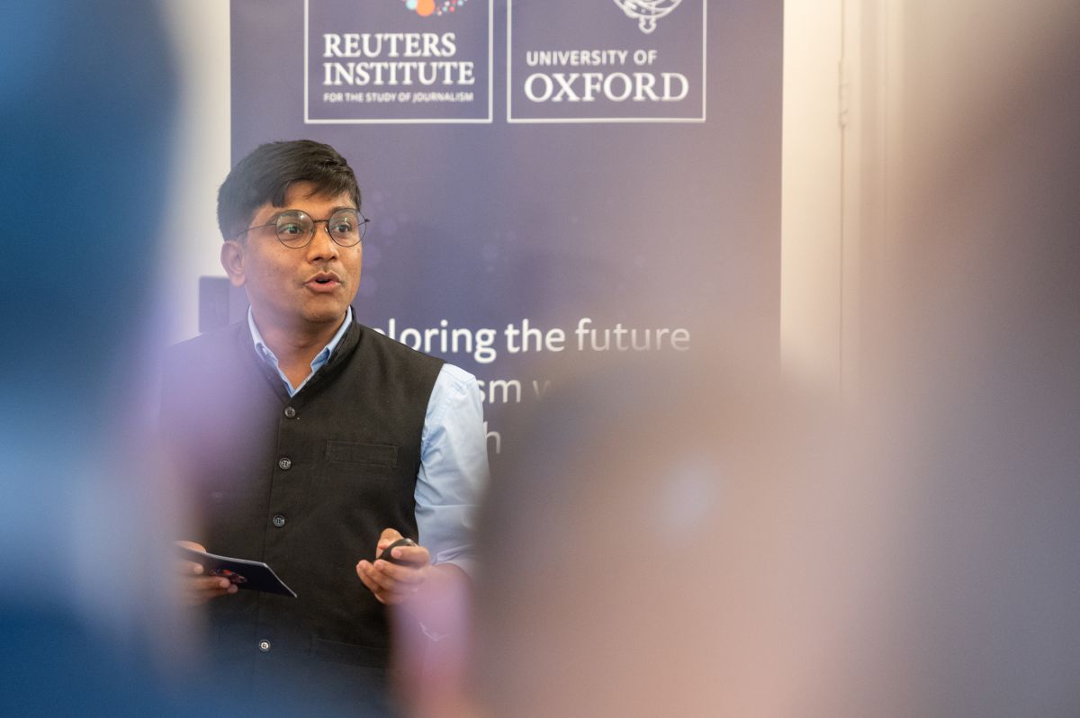 Ganesh Pol speaks while presenting at the Reuters Institute, standing in front of a backdrop displaying the Reuters Institute and University of Oxford logos. He wears glasses, a black waistcoat, and holds a presentation remote and notecards.