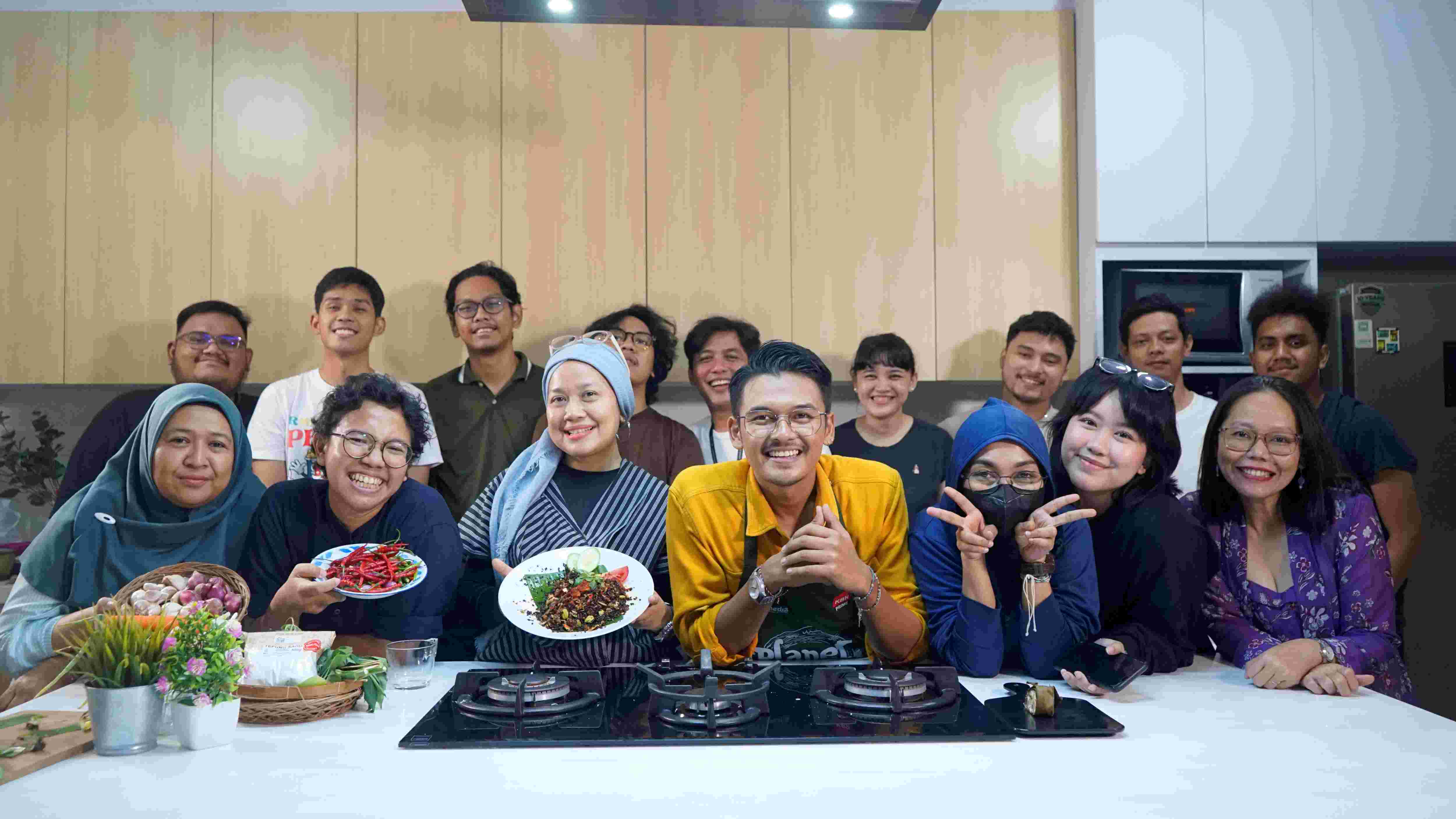 A group of people huddle in a kitchen posing for a picture. Three people on the left-hand side are holding plates of food.