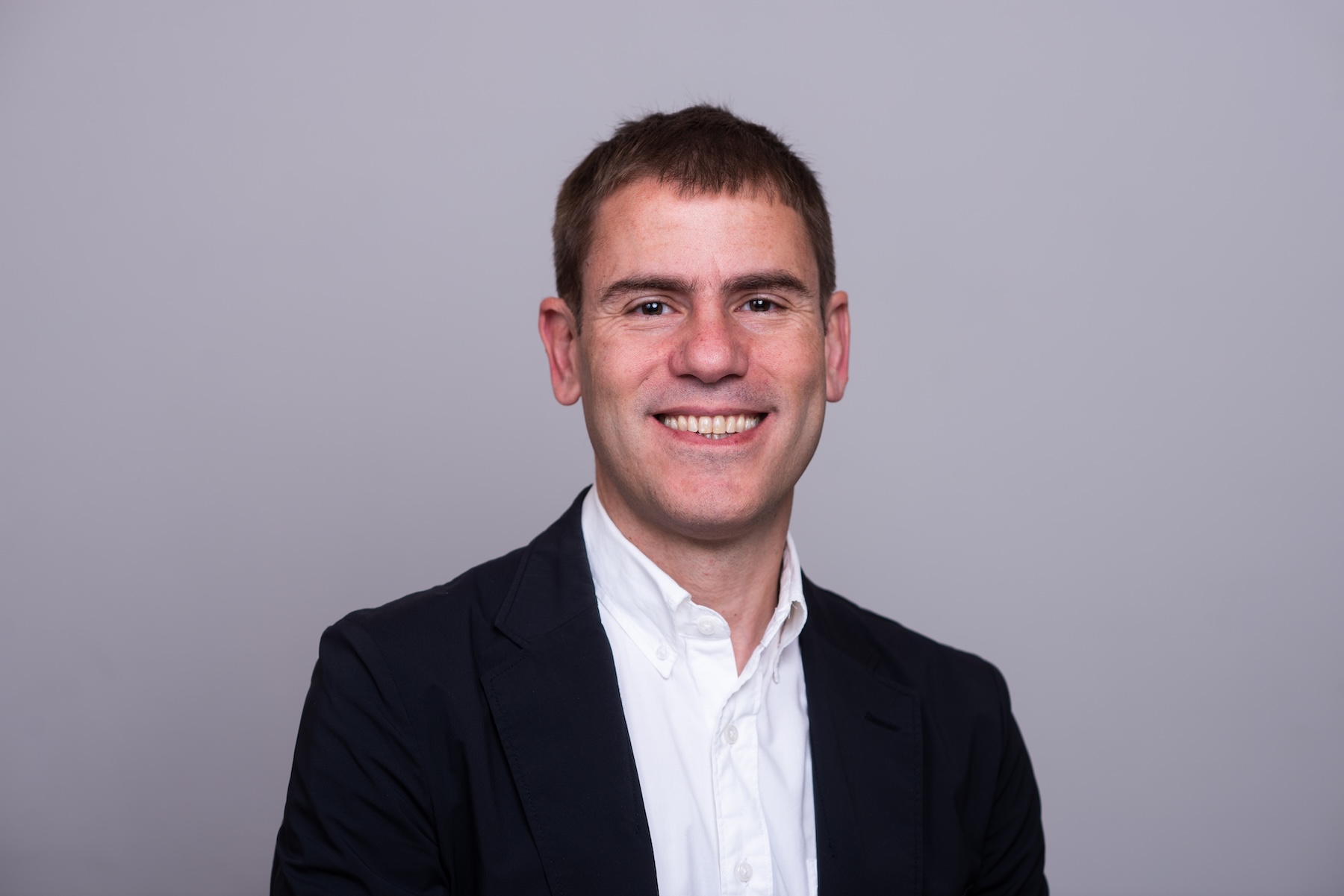 Smiling man in a white button-up shirt and dark blazer, facing the camera against a plain light gray background