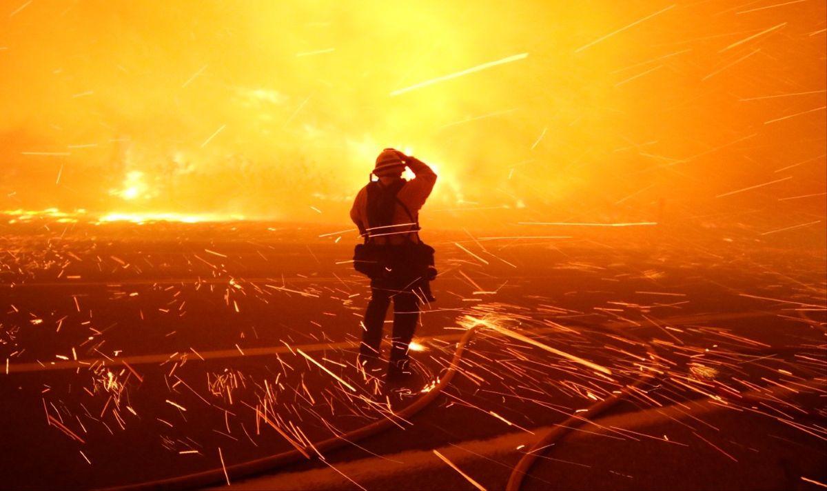 A firefighter stands on a road facing an intense wildfire, silhouetted against a wall of bright orange flames and smoke. Sparks and embers fly across the dark pavement as the blaze rages in the background.