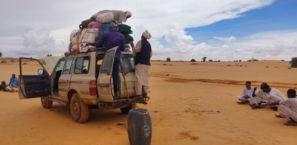 An offroad vehicle idle in the desert. One of the front and back doors are open and two men perch on the back step, arranging bundles wrapped in fabric that are tied to the top of the car. A third man sits on top of the bundles and appears to be securing the rope. Two small groups of people, mostly men, sit on the orange sand around the car.