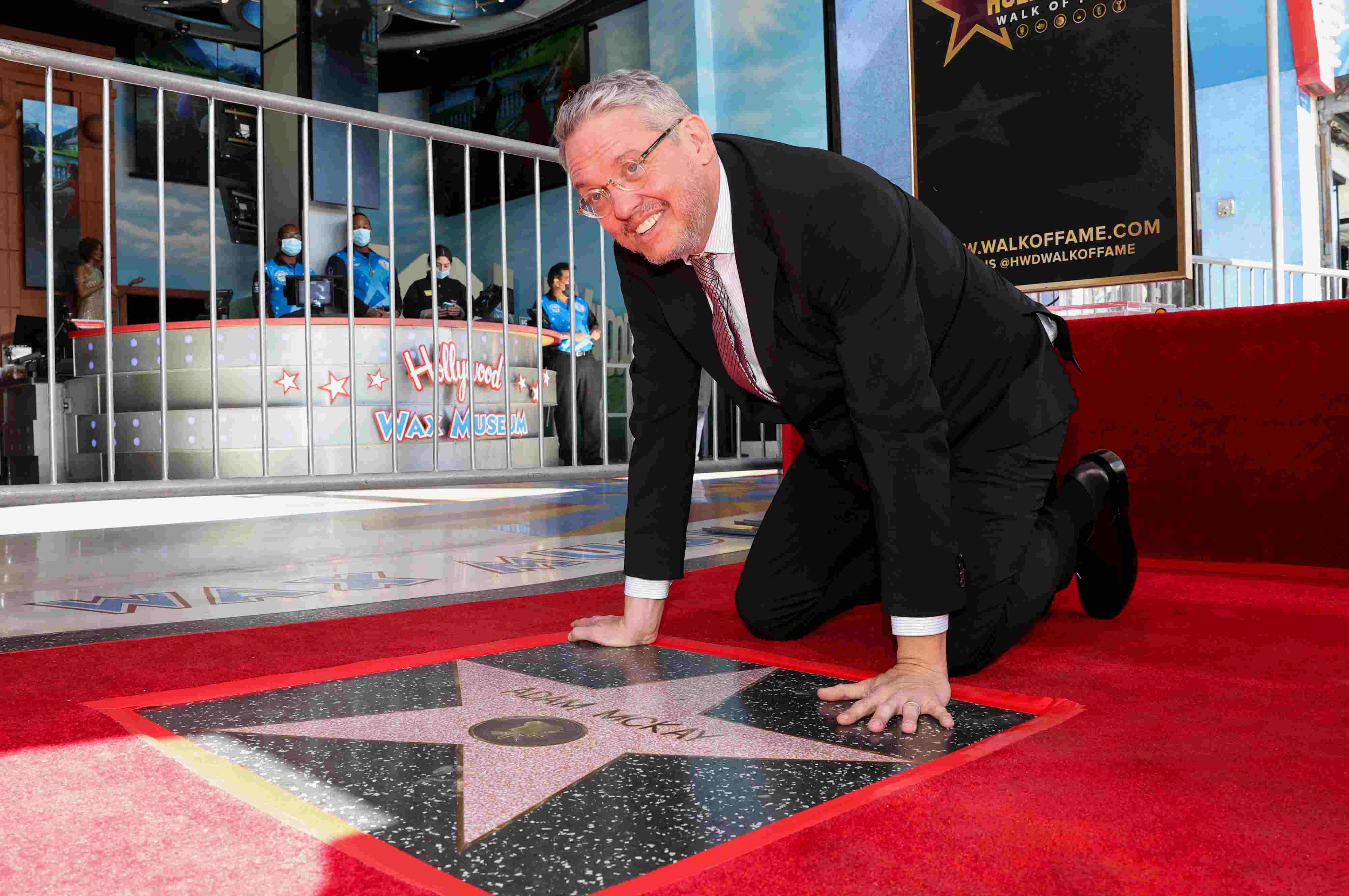 A grinning man kneels over a star shape in the pavement with the name 'Adam McKay' engraved on it.
