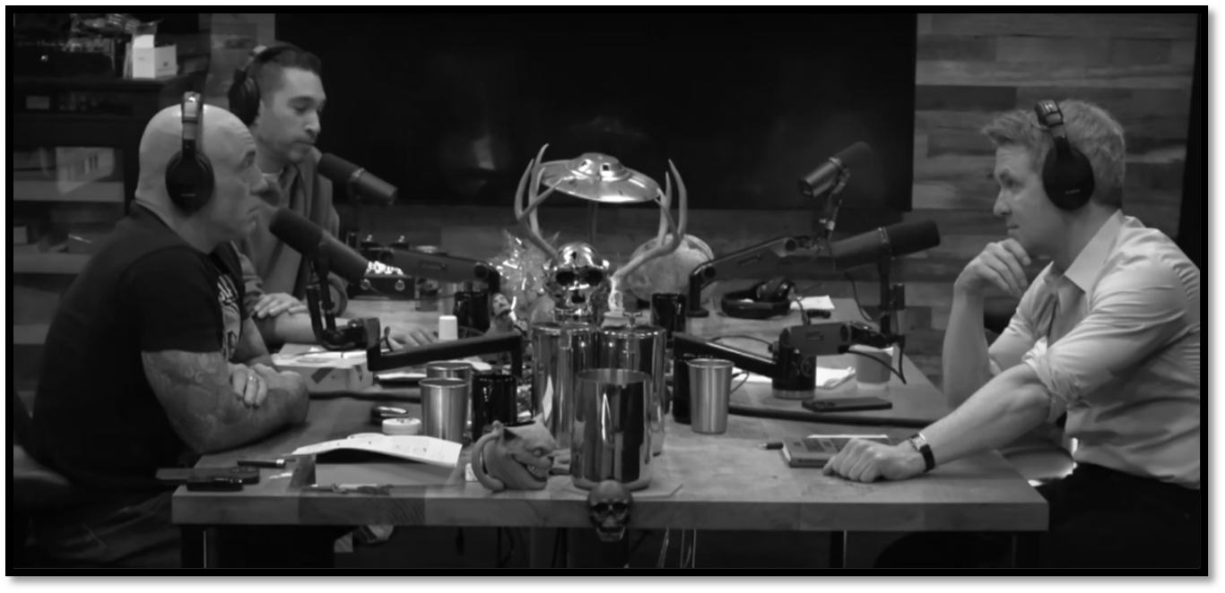 Joe Rogan sits at a podcast table with guests Dave Smith and Douglas Murray as they converse into microphones, engaged in a discussion about trust during an episode of The Joe Rogan Experience.