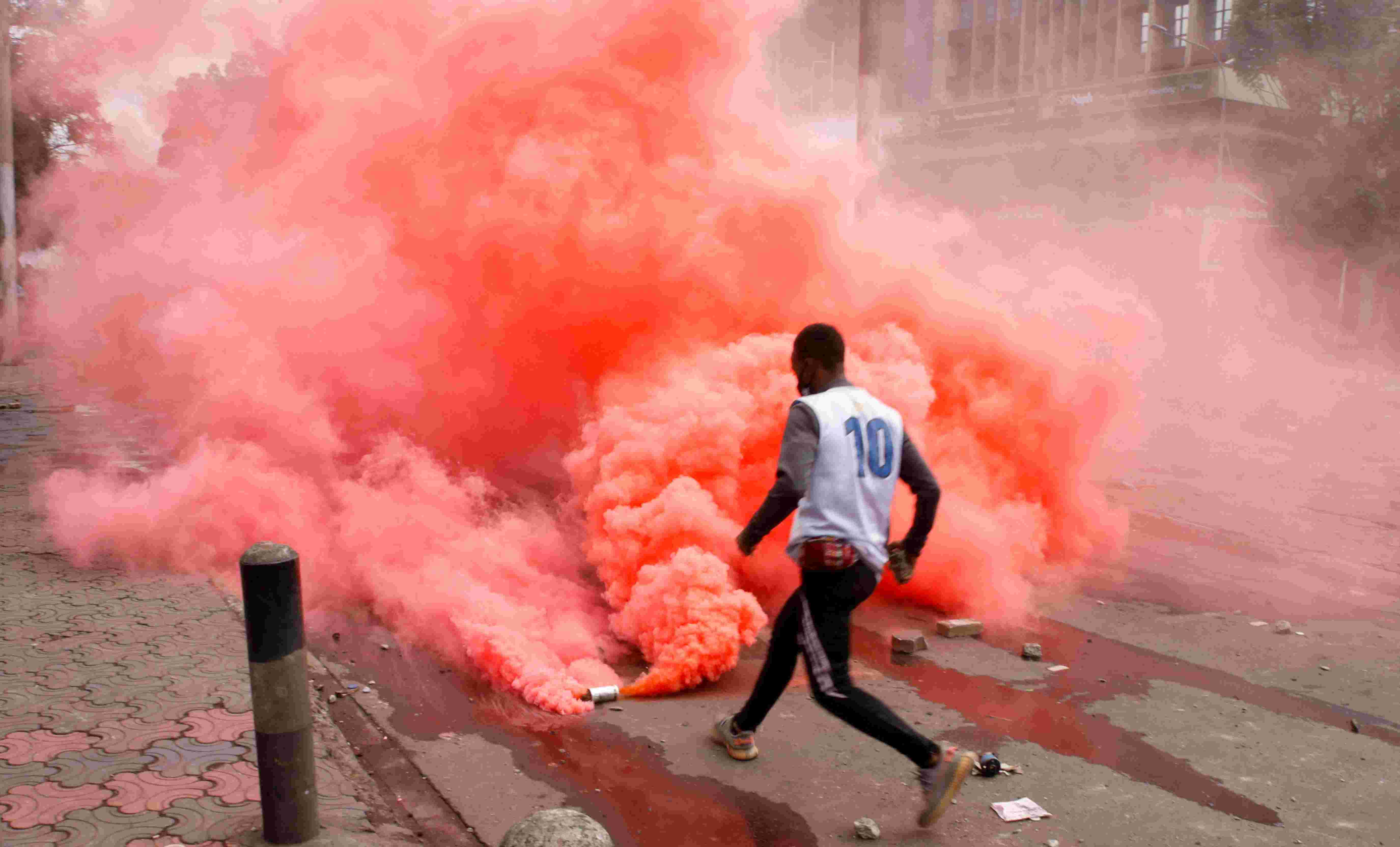 A man runs towards a canister, out of which a large orange cloud is billowing out into the street.