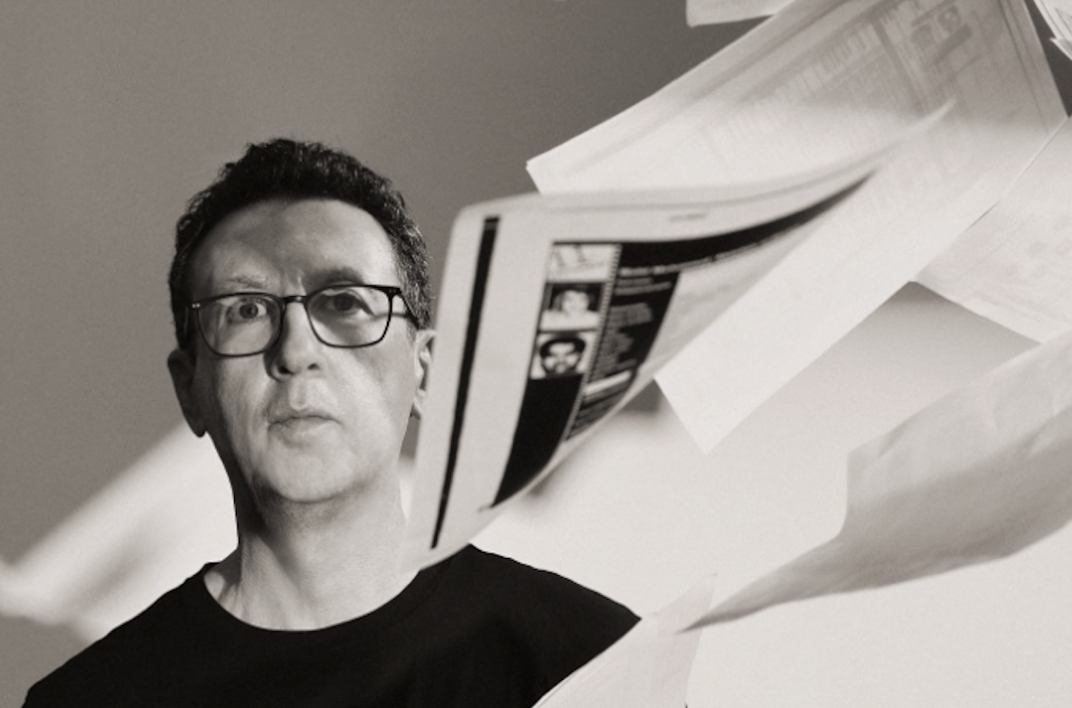 A grayscale photo of a man in a black t-shirt looking serious, standing as a flurry of printouts fall around him.