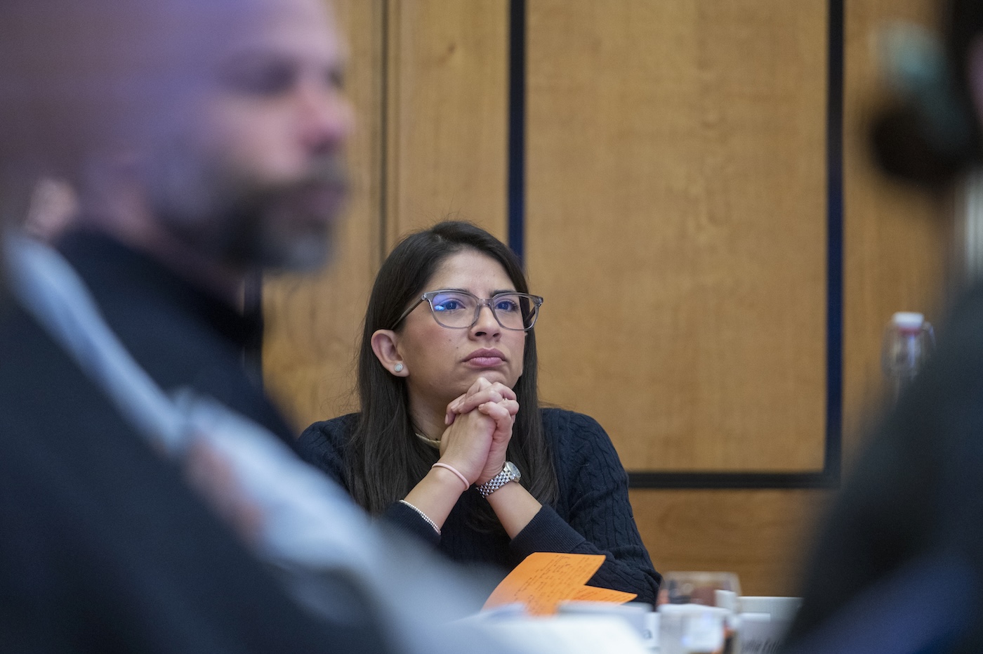 A woman with glasses sits at a table with her hands clasped, looking forward with a focused expression. Other participants are partially visible in the foreground.