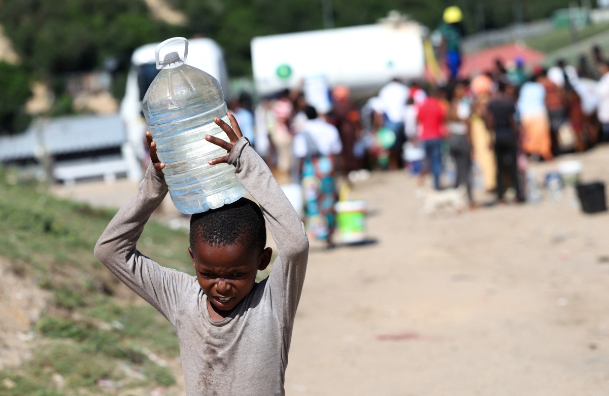 A boy carries water balanced on his head. In the background you can see a crowd of people gathered around a truck.