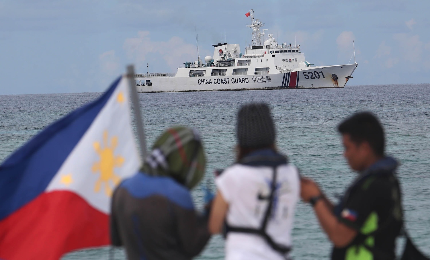 A China Coast Guard ship with the number 5201 is seen in the distance on a calm sea. In the blurred foreground, three people stand near a Philippine flag, observing or filming the vessel.