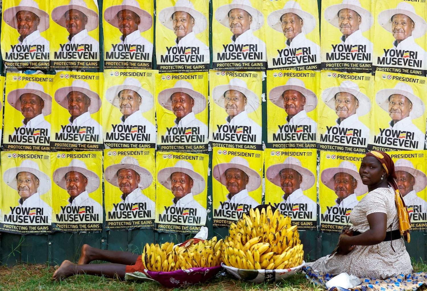 A woman and a child sit on the ground in front of a wall covered with campaign posters for Yoweri Museveni, with large bunches of yellow bananas in the foreground.