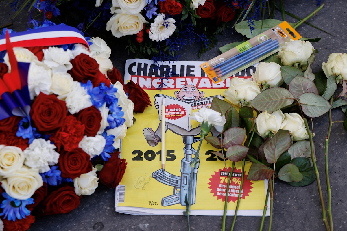 A memorial display features a copy of the French satirical magazine Charlie Hebdo, surrounded by red, white, and blue flower bouquets, loose white roses, and several pencils. The magazine cover depicts a cartoon of a man reading, seated atop the barrel of an upright rifle.