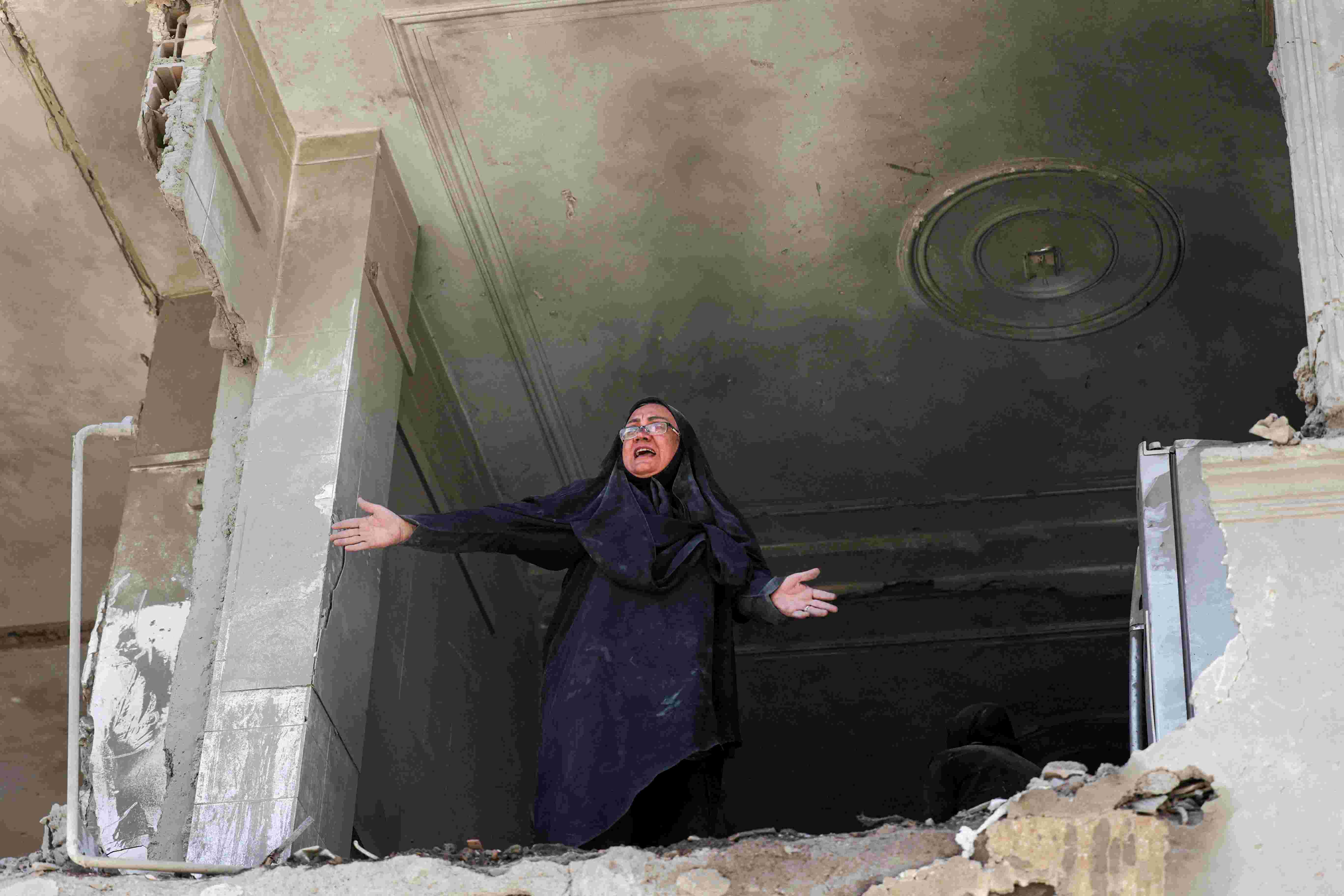 A woman gestures with her arms, looking out of a room where the exterior wall has collapsed, allowing people to see inside the apartment she is in.  