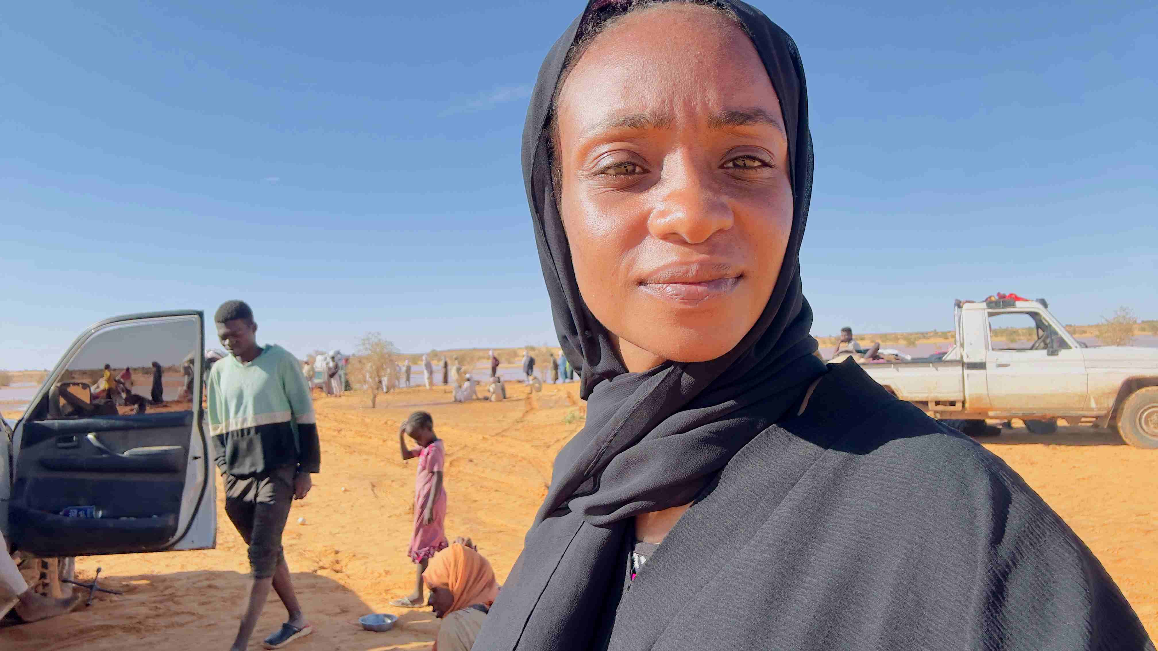 A close-up photo of a woman dressed in black. Behind her is a desert landscape with two idle vehicles and people standing or sitting around on the sand, including a child.