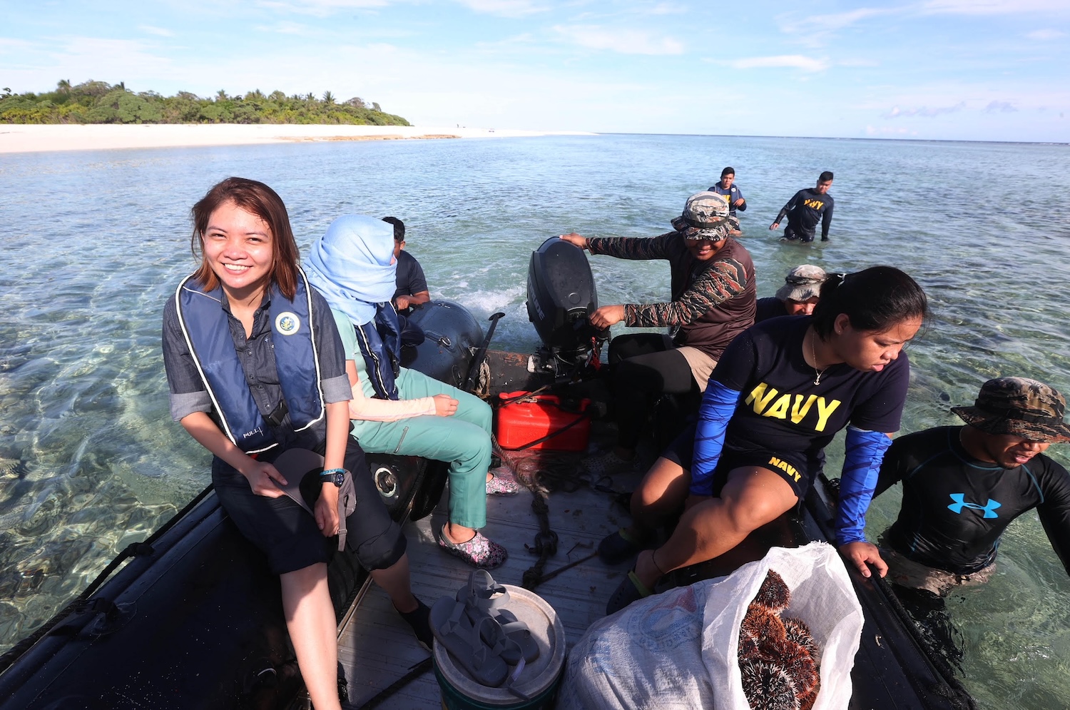 A female journalists looks at the camera while on a boat in the sea, near the seashore.