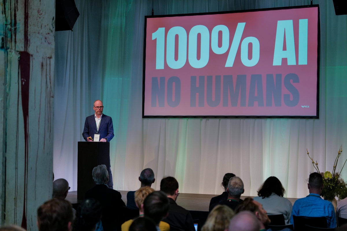 A man stands at a lectern in front of a room of people. Behind him on a large screen are the words: "100% AI NO HUMANS"