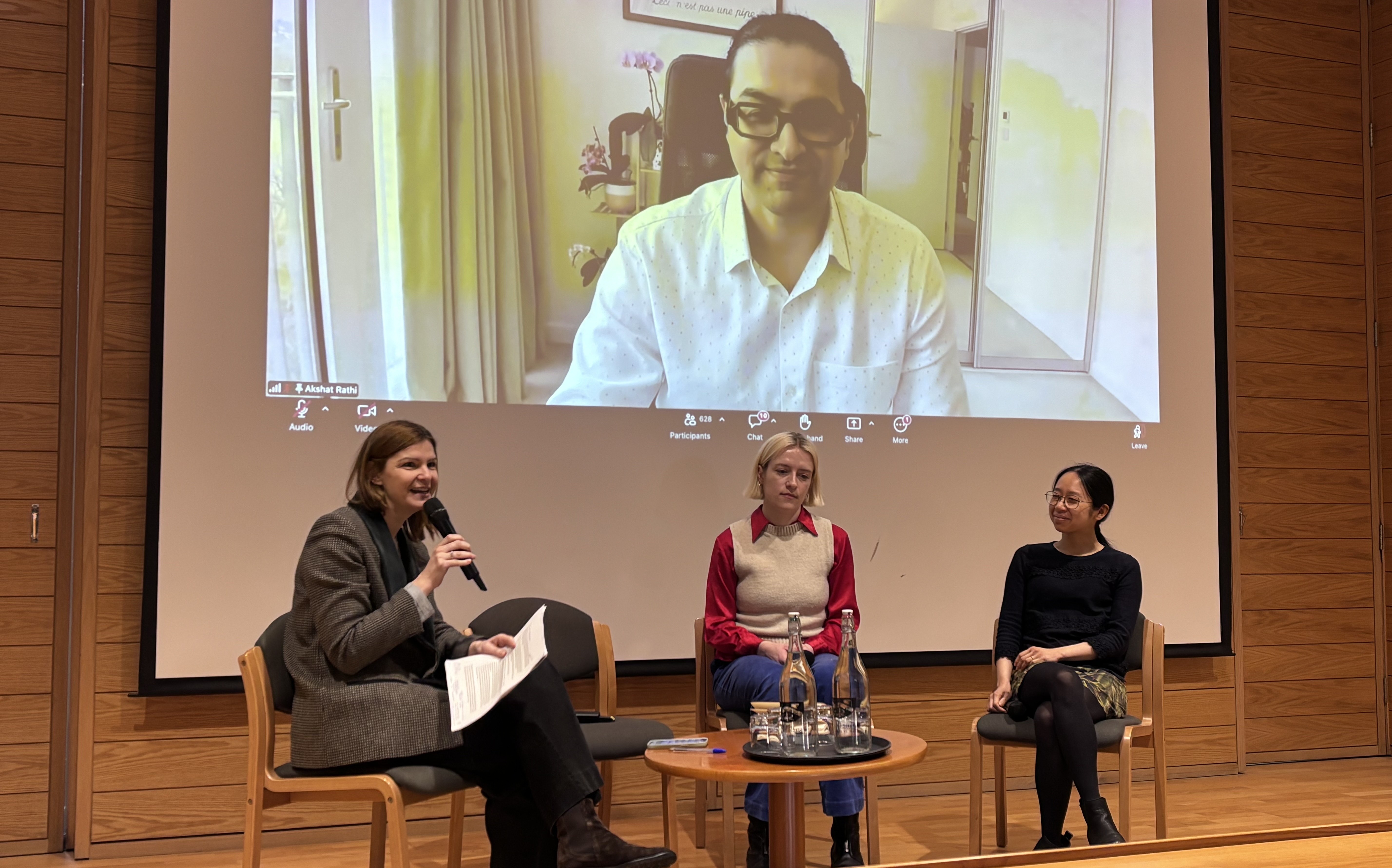 Three women sit on chairs before a small table with water bottles and glasses. One of them is speaking into a microphone and holding a piece of paper with writing on it. Behind them is the image of a man on a Zoom call projected onto a large screen.