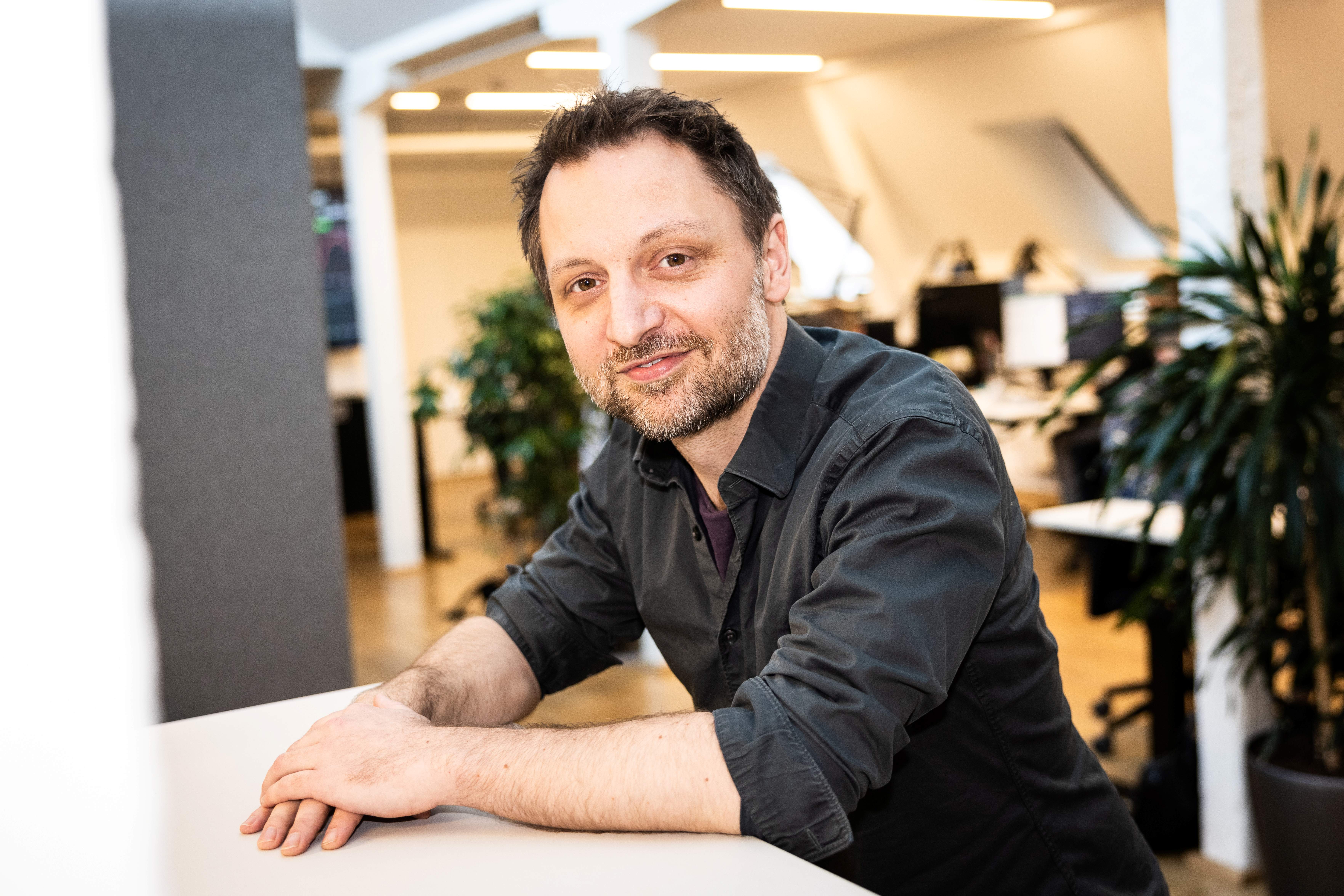 A photograph of Kasper Lindskow leaning on a table with both hands on the tabletop, smiling.