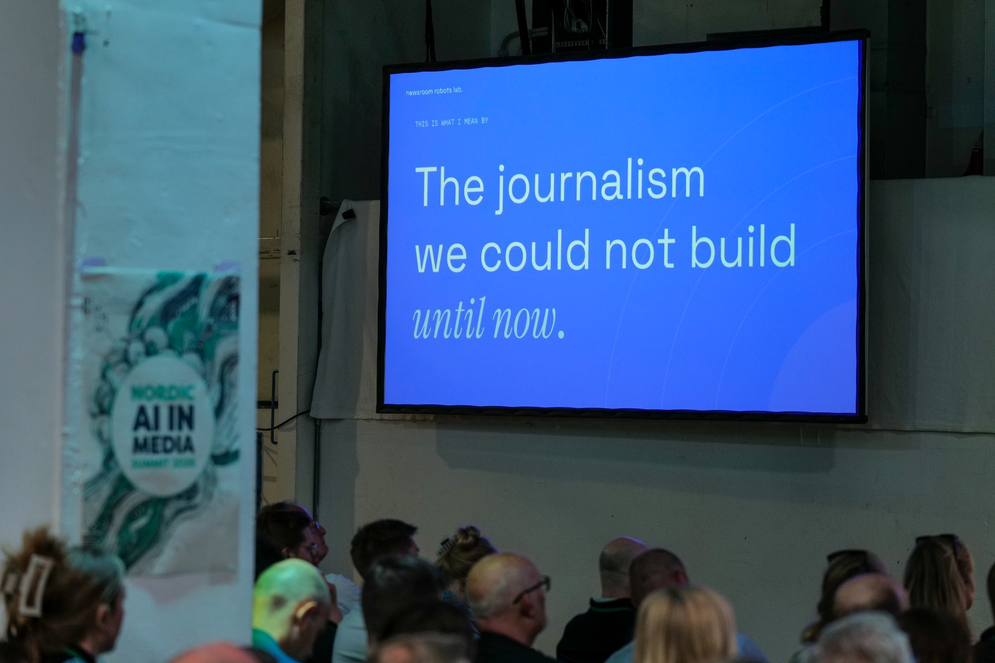 A photograph of a room focused on a large blue screen in front of rows of seated people. The text on the screen reads: "The journalism we could not build until now."