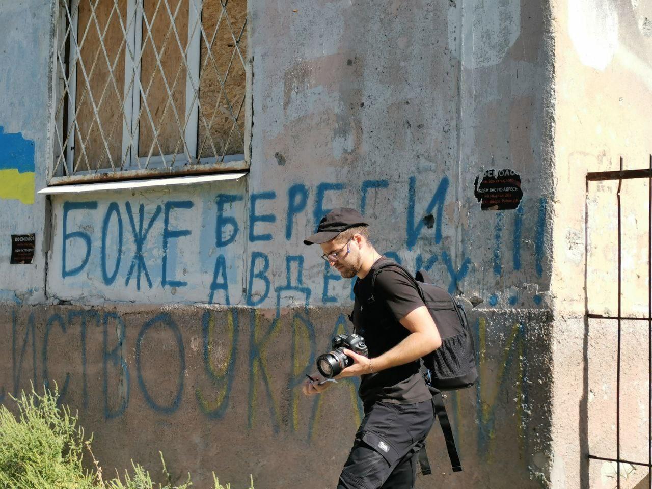 A man walks carrying a camera, looking at the floor. Behind him is a building with graffiti in Ukrainian, including a Ukrainian flag.