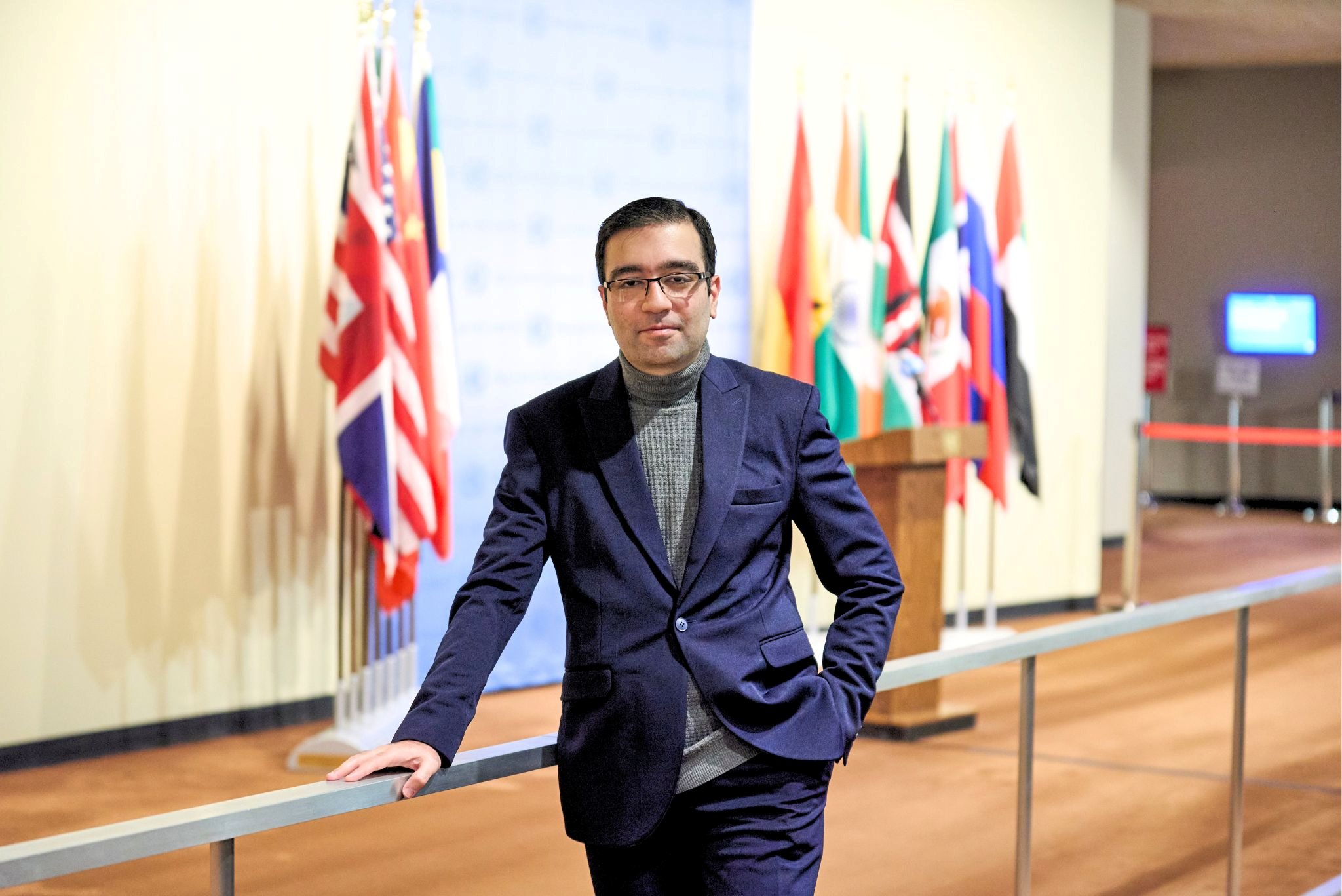 A man wearing a gray turtleneck and a navy suit poses in front of several flags belonging to different nations.