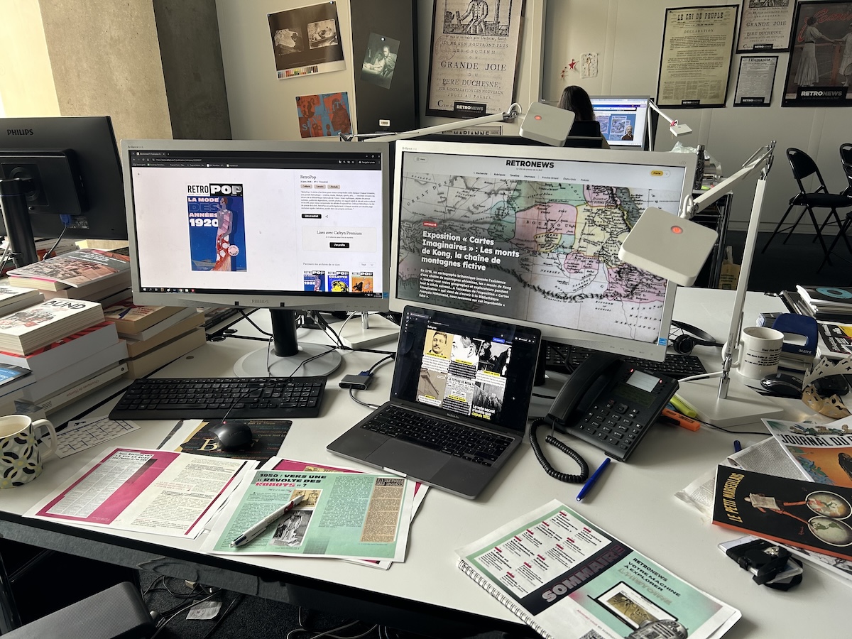 A busy office desk features two desktop monitors and a laptop displaying the RetroNews website and digital archives. The workspace is cluttered with stacks of books, printed historical articles, a desk phone, and a mug, with vintage-style newspaper posters visible on the wall in the background.