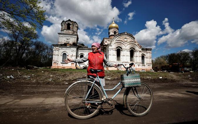 An elderly woman holding a bicycle gestures while speaking in front of a ruined church.