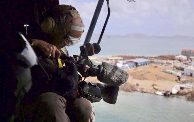 Maxine holds a camera while shooting an aerial shot from a helicopter above an island.