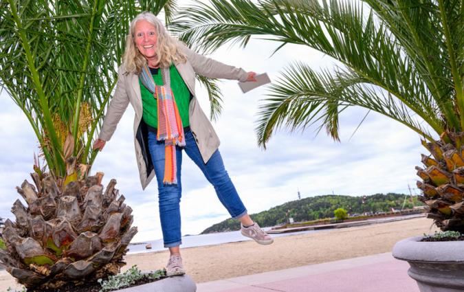 A blonde woman in jeans, green t-shirt and colourful scarf balances on a palm tree plant pot on an Oslo beachfront.