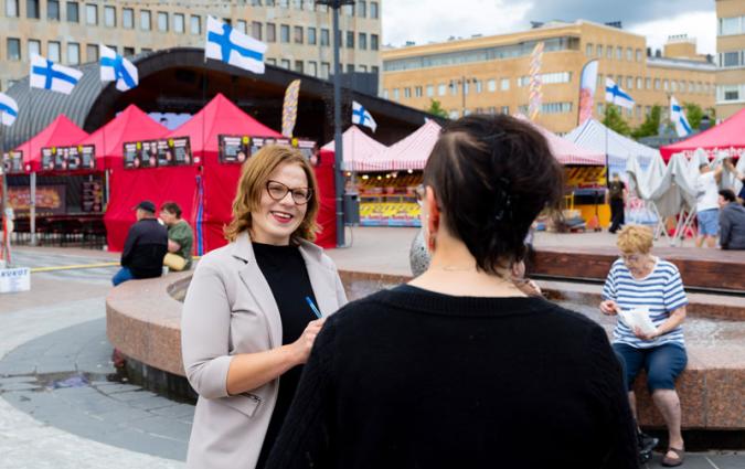 A market square with Finnish flags and red market stalls. A woman with blonde hair is interviewing a woman dressed in black.. 