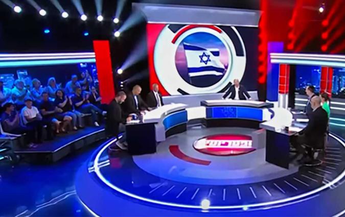 A host and 6 guests sit around a circular table in a TV studio with a prominent Israeli flag on a screen behind them