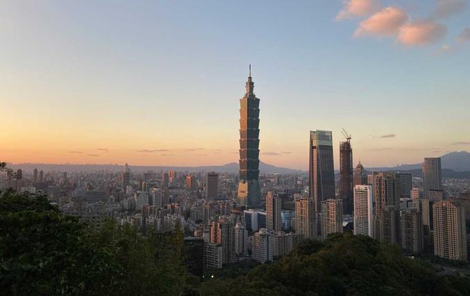 A panoramic view of Taipei at sunset, featuring Taipei 101 towering over the city's skyline. The sky transitions from soft blue to warm orange hues, with scattered clouds reflecting the evening light.