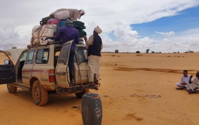 An offroad vehicle idle in the desert. One of the front and back doors are open and two men perch on the back step, arranging bundles wrapped in fabric that are tied to the top of the car. A third man sits on top of the bundles and appears to be securing the rope. Two small groups of people, mostly men, sit on the orange sand around the car.
