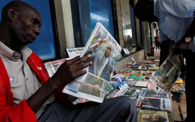 A man sits on a street pavement looking at a newspaper featuring a prominent photo of former Kenyan President Daniel arap Moi on the front page, while surrounded by stacks of magazines and books on display.