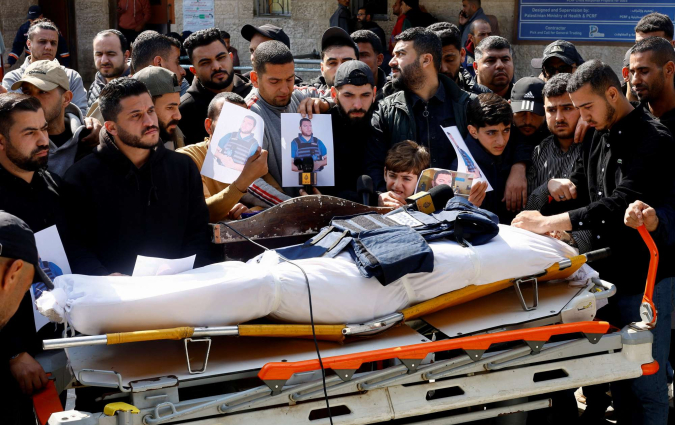 A body wrapped in white bandages lies on a stretcher, a press vest and microphone laid on top, as mourners gather and hold up photos of a man in a press vest.