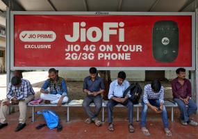 Commuters use their mobile phones as they wait at a bus stop with an advertisement of Reliance Industries' Jio telecoms unit, in Mumbai, India July 10, 2017. REUTERS/Shailesh Andrade