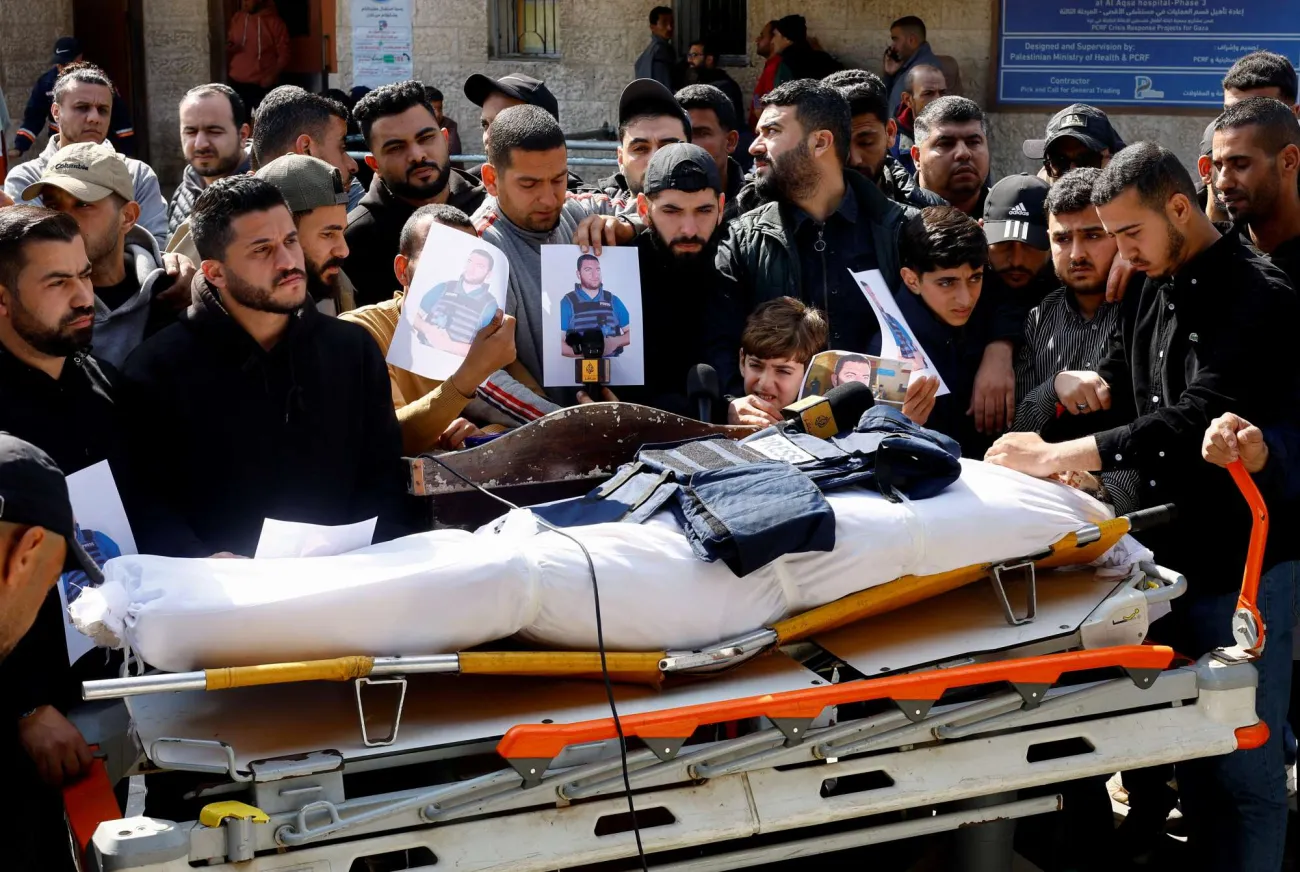 A body wrapped in white bandages lies on a stretcher, a press vest and microphone laid on top, as mourners gather and hold up photos of a man in a press vest.