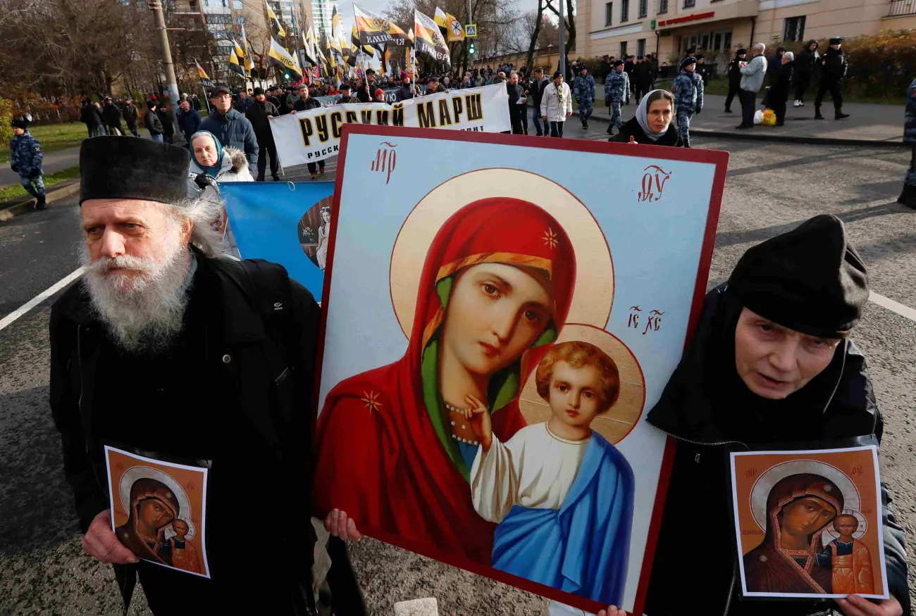 A man and woman in black Russian Orthodox clothing hold up Orthodox religious iconography in the street. Behind them are other people holding up banners and signs.