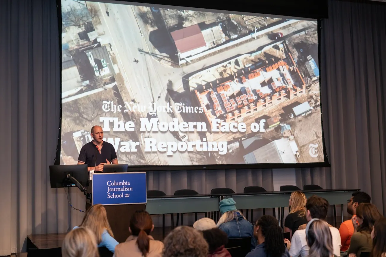 Speaker at a Columbia Journalism School event presenting “The Modern Face of War Reporting” to an audience, with the title displayed on a large screen behind them.