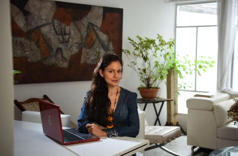 A woman with long brown hair sits at a white table with her computer, abstract art on the wall behind her